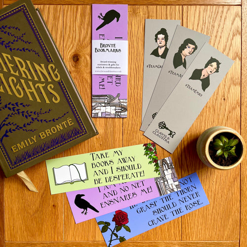 Photo of bookmarks laid out next to a fancy hardback edition of Wuthering Heights and a tiny plant. The bookmarks are spread out and feature quotes from the Brontes on one side alongside images of ivy, a book, a crow, a ruin and a rose. On the other, the portraits of the sisters with a Goth makeover, and 