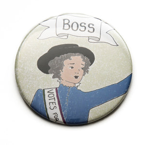 Photo of a pocket mirror showing the design of a Suffragette with her arm raised and the caption above "Boss" on a banner. She is wearing blue.