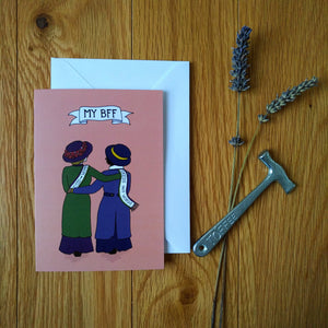 Photo of a greetings card depicting two suffragettes arm in arm from behind, one with a "Votes for Women" sash and green coat, the other with a "Deeds Not Words" scarf and blue coat. Above is a banner reading "My BFF" and the background is a peach colour. Also pictured besides the card & envelope are a toffee hammer and a spring of lavender.