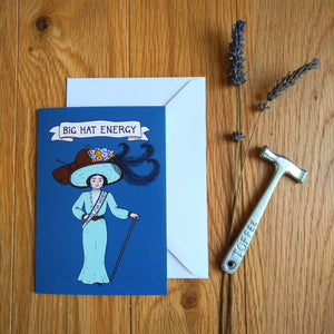 Photo of a greetings card depicting a very elegant suffragettes with a comically gigantic hat featuring a pile of flowers, massive feathers and an epic bow. Above is a banner reading "Big Hat Energy" and the background is a teal colour. Also pictured besides the card & envelope are a toffee hammer and a spring of lavender.