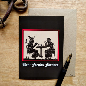Photo of a greetings card featuring an early modern woodcut image of two witch-like women socializing over a small table. Underneath is the caption 'Best Fiends Forever' and the woodcut is surrounded by a scarlet border and the background is black. Along with the brown envelope, also pictured are a black fountain pen and a gnarled branch.