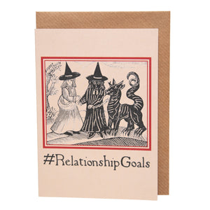 A buff coloured card featuring a mediaeval woodcut artwork depicting two witches, one of which is holding a pet demon. Beneath it the caption reads #RelationshipGoals. Pictured with a brown envelope.