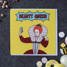 Load image into Gallery viewer, Photo of a large square yellow greetings card featuring a comic book style illustration of Queen Elizabeth I taking a selfie, with &#39;Beauty Queen&#39; written in a flash above her. Dotted around the card &amp; white envelope are pearls like the ones she wears, and sweets in yellows and whites to match the card colour.