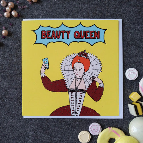 Photo of a large square yellow greetings card featuring a comic book style illustration of Queen Elizabeth I taking a selfie, with 'Beauty Queen' written in a flash above her. Dotted around the card & white envelope are pearls like the ones she wears, and sweets in yellows and whites to match the card colour.