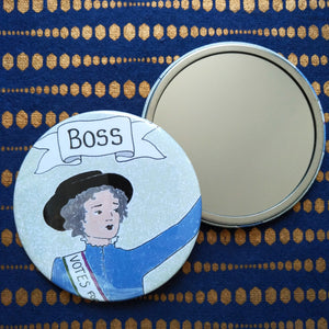 Photo of a pocket mirror on its back with the mirror showing, and another on its front showing the design of a Suffragette with her arm raised and the caption above "Boss" on a banner. She is wearing blue. Also the background is blue with a gold pattern.