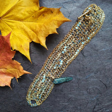 Load image into Gallery viewer, Photo of a crocheted wire cuff laid flat, studded with turquoise coloured beads and with a teal coloured large pointed bead for a clasp pin. Also pictured are the edges of golden coloured leaves.