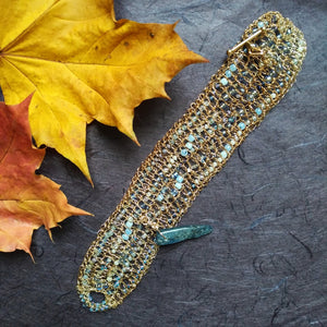Photo of a crocheted wire cuff laid flat, studded with turquoise coloured beads and with a teal coloured large pointed bead for a clasp pin. Also pictured are the edges of golden coloured leaves.