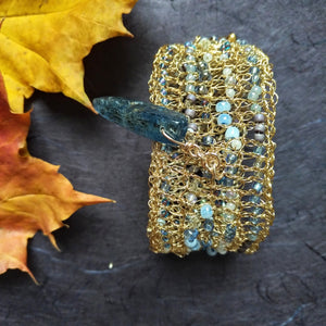 Photo of a crocheted wire cuff, studded with turquoise coloured beads and held together with a teal coloured large pointed bead for a clasp pin. Also pictured are the edges of golden coloured leaves.