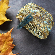 Load image into Gallery viewer, Photo of a crocheted wire cuff, studded with turquoise coloured beads and held together with a teal coloured large pointed bead for a clasp pin. Also pictured are the edges of golden coloured leaves.