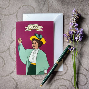 Photo of a greetings card depicting a woman in Edwardian dress with her arm raised and her mouth open as if exhorting her troops. Above is a banner reading "Boss" and the background is a plum colour. Also pictured besides the card & envelope are a green fountain pen and a spring of lavender.