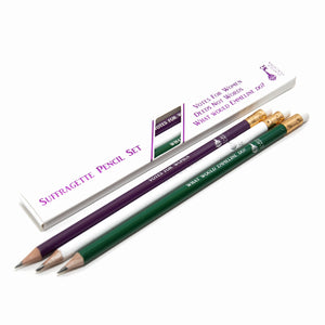 Set of three graphite pencils pictured next to handmade paper and a sprig of lavender. The pencils are painted purple, green & white, featuring slogans "Votes for Women", "Deeds Not Words" and "What would Emmeline do?". They are presented in a box.