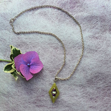 Load image into Gallery viewer, Photo depicting a silver coloured necklace with a moss green diamond pendant, within which are pearl coloured and dark purple beads. A purple flower sits to the side.