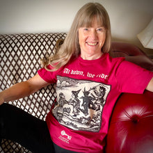 Load image into Gallery viewer, Photograph of a t-shirt modelled by a smiling white-skinned woman with long silvery brown hair. The cherry red coloured t-shirt features a monochrome mediaeval woodcut design depicting a woman addressing witches and a demon each mounted on broomsticks. Above in Old English text are the words &quot;At dawn we ride&quot;.