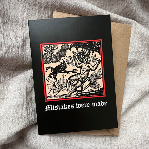 A black coloured card featuring a mediaeval woodcut artwork depicting a person holding up their hands as they fall into the jaws of hell, tormented by a devil with a pitchfork. Beneath it the caption reads Mistakes were made. Pictured with a brown envelope, and resting on a beige fabric.