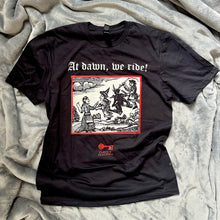 Load image into Gallery viewer, Photograph of a t-shirt laid out on a pale background. The black coloured t-shirt features a monochrome mediaeval woodcut design with red border depicting a woman addressing witches and a demon each mounted on broomsticks. Above in Old English text are the words "At dawn we ride".