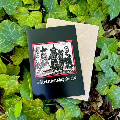 A black coloured card featuring a mediaeval woodcut artwork depicting two witches, one of which is holding a pet demon. Beneath it the caption reads #RelationshipGoals. Pictured with a brown envelope, and resting on a bed of ivy.