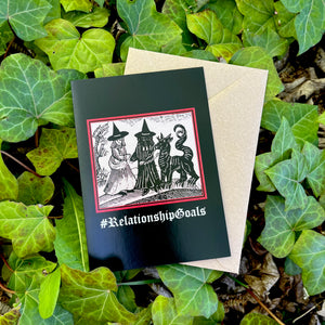 A black coloured card featuring a mediaeval woodcut artwork depicting two witches, one of which is holding a pet demon. Beneath it the caption reads #RelationshipGoals. Pictured with a brown envelope, and resting on a bed of ivy.