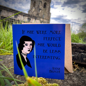 Photo of a blue greetings card on a mossy stone with an old church in the distance. The card depicts the quote 'If she were more perfect, she would be less interesting' by Anne Bronte, with a portrait of her as a Goth.
