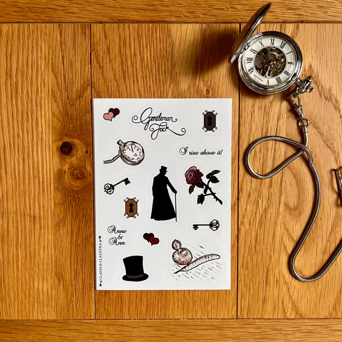 Photo of a glossy A6 sheet with Gentleman Jack stickers: interlocking pink and black hearts; “Gentleman Jack” in stylised writing; a dark keyhole; a rose with thorns; a pocket watch and chain; an ornate iron key; a light keyhole; a silhouette of Anne Lister with a top hat and cane; an ornate iron key with a different design, interlocking burgundy and black hearts; “I rise above it!” In stylised writing; “Anne & Ann” in stylised writing; a top hat; and a quill and ink pot. A silver ink pot is beside it.