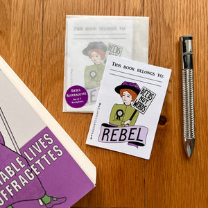 Photograph of a rectangular sticker featuring a suffragette with red hair and a green outfit wearing a rosette and holding a placard reading “Votes For Women”. Below her is a banner reading “Rebel and above other words “This book belongs to:…” – with room to write your name. Also in the photo is a packet of these bookplate stickers, Diane Atkinson’s book Rise Up, Women, and a silver pen.