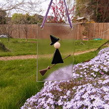 Load image into Gallery viewer, Photo of glasswork which depicts a witch in a minimalist style, with a black dress and black hat, flying on a broomstick. The artwork hands from a purple thread outside, with a fence and grass and flowers in the background.