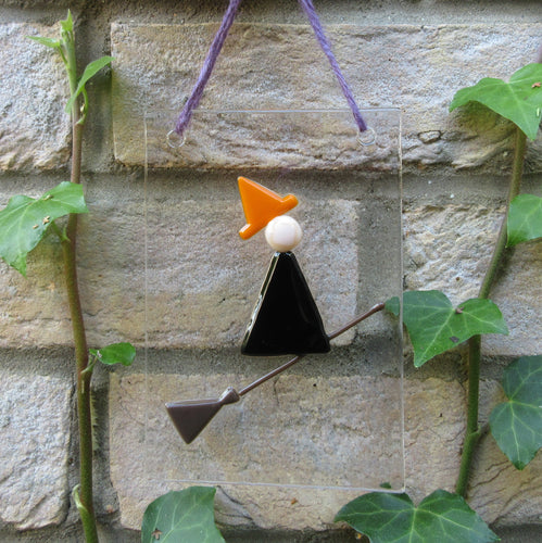 Photo of glasswork which depicts a witch in a minimalist style, with a black dress and orange hat, flying on a broomstick. The artwork hands from a purple thread against a brick wall with ivy.