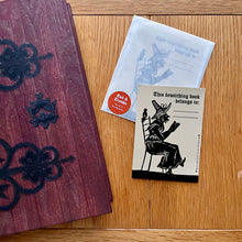 Load image into Gallery viewer, Photograph of a rectangular sticker featuring a Mediaeval woodcut image of a woman sitting on a chair and reading a book, wearing a pointed hat, with a bird perched behind her. Above other words “This bewitching book belongs to:…” – with room to write your name. Also in the photo is a packet of these bookplate stickers and a hardback notebook which looks like a large oak door.