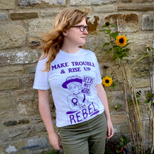Load image into Gallery viewer, Photo of a model wearing a white t-shirt with an illustration and text, all in purple. The image is of a suffragette wearing a hat with flowers, a rosette, and holding a sign that says “Deeds Not Words”. Beneath her is a banner that says “Rebel”. Above is the caption “Make Trouble &amp; Rise Up”. The model has long ginger hair and is looking to the side smiling, standing next to a wall with two sunflowers.