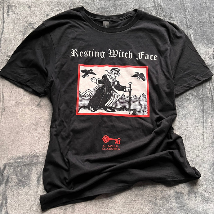 Photo of a black t-shirt featuring a black-and-white mediaeval woodcut artwork on a black background and framed in red, depicting a stereotypical crone with warts and a large nose in a black dress, holding a cane and riding a raft on water. Two black birds hover in the sky. Above it the caption reads 'Resting Witch Face' in a white Gothic typeface. Underneath is the Clavis & Claustra logo in red.