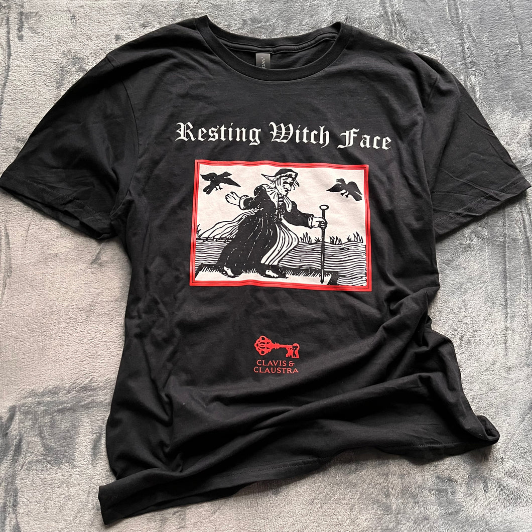 Photo of a black t-shirt featuring a black-and-white mediaeval woodcut artwork on a black background and framed in red, depicting a stereotypical crone with warts and a large nose in a black dress, holding a cane and riding a raft on water. Two black birds hover in the sky. Above it the caption reads 'Resting Witch Face' in a white Gothic typeface. Underneath is the Clavis & Claustra logo in red.