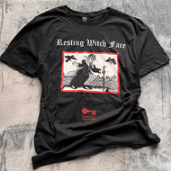 Photo of a black t-shirt featuring a black-and-white mediaeval woodcut artwork on a black background and framed in red, depicting a stereotypical crone with warts and a large nose in a black dress, holding a cane and riding a raft on water. Two black birds hover in the sky. Above it the caption reads 'Resting Witch Face' in a white Gothic typeface. Underneath is the Clavis & Claustra logo in red.