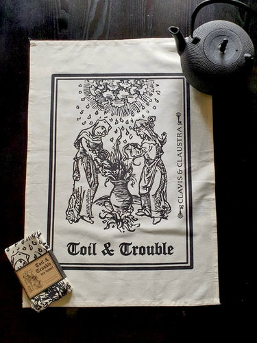 Photo of a natural cotton coloured tea towel on a black background, next to a black teapot and a folded up separate tea towel bound in brown cardboard and reading “Toil & Trouble”. The tea towel shows a mediaeval woodcut of two women around a bubbling cauldron as the clouds rain overhead. The caption reads 'Toil & Trouble' in a black Gothic typeface.