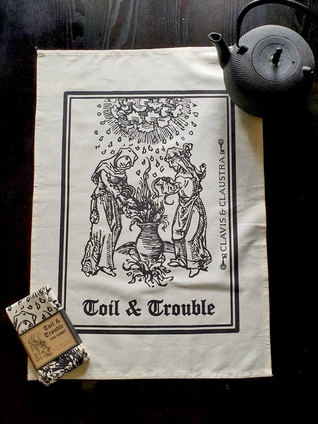 Photo of a natural cotton coloured tea towel on a black background, next to a black teapot and a folded up separate tea towel bound in brown cardboard and reading “Toil & Trouble”. The tea towel shows a mediaeval woodcut of two women around a bubbling cauldron as the clouds rain overhead. The caption reads 'Toil & Trouble' in a black Gothic typeface.
