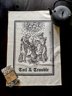 Photo of a natural cotton coloured tea towel on a black background, next to a black teapot and a folded up separate tea towel bound in brown cardboard and reading “Toil & Trouble”. The tea towel shows a mediaeval woodcut of two women around a bubbling cauldron as the clouds rain overhead. The caption reads 'Toil & Trouble' in a black Gothic typeface.