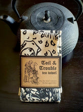 Load image into Gallery viewer, Photo of a folded up natural cotton coloured tea towel bound in brown cardboard next to a black teapot. Part of the mediaeval woodcut showing a woman’s face is visible. The caption reads &#39;Toil &amp; Trouble tea towel’ and ‘For the chief sorcerer or her apprentice’ in a black Gothic typeface.