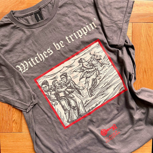 Photograph of a t-shirt laid out on a wooden table. The charcoal coloured t-shirt features a mediaeval woodcut design depicting a group of people talking and one woman riding off on a winged demon. Above in Old English text are the words 