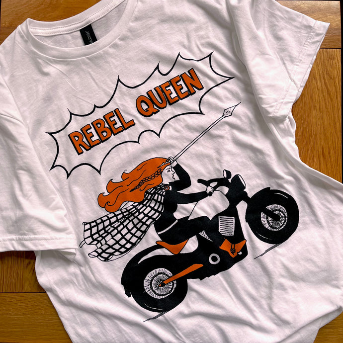 Photograph of a t-shirt laid out on a wooden table. The white coloured t-shirt features an illustration in black and flaming orange of the Roman-era Celtic icon Boudicca riding a motorbike and brandishing a spear, underneath the text 