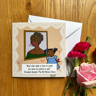 Greeting card with cartoon illustration of an older woman’s portrait and a young child, where the child has drawn in crayon on the woman’s portrait; the card surrounded by flowers on a wooden surface.