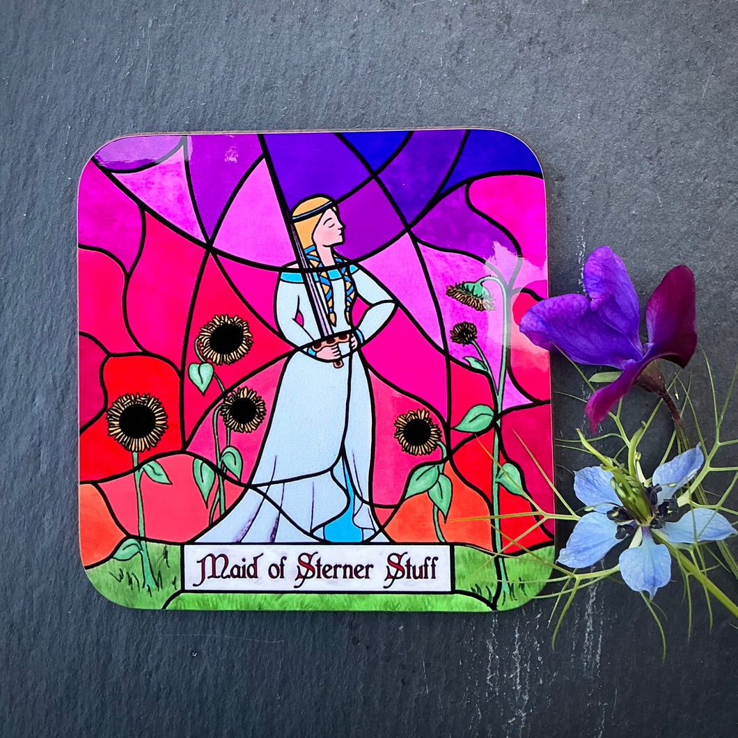 Photo of drinks coaster depicting a stained glass window set in a stone wall. The glass design shows a woman in mediaeval dress casually wielding a sword and surrounded by sunflowers against a sunset sky. The caption reads 'Maid of Sterner Stuff'. The coaster is with two blue/purple flowers on a slate background.