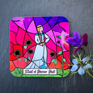 Photo of drinks coaster depicting a stained glass window set in a stone wall. The glass design shows a woman in mediaeval dress casually wielding a sword and surrounded by sunflowers against a sunset sky. The caption reads 'Maid of Sterner Stuff'. The coaster is with two blue/purple flowers on a slate background.