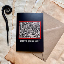 Load image into Gallery viewer, Photo of a black coloured card featuring a mediaeval woodcut artwork depicting witches approaching a demonic figure with a goat’s head and wings who sits on a throne and beckons to them. Beneath it the caption reads &#39;Haters gonna hate!&#39;. Pictured with a brown envelope and black letter opener with a curved, spiral end.
