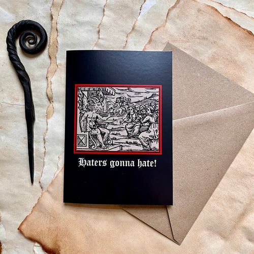 Photo of a black coloured card featuring a mediaeval woodcut artwork depicting witches approaching a demonic figure with a goat’s head and wings who sits on a throne and beckons to them. Beneath it the caption reads 'Haters gonna hate!'. Pictured with a brown envelope and black letter opener with a curved, spiral end.