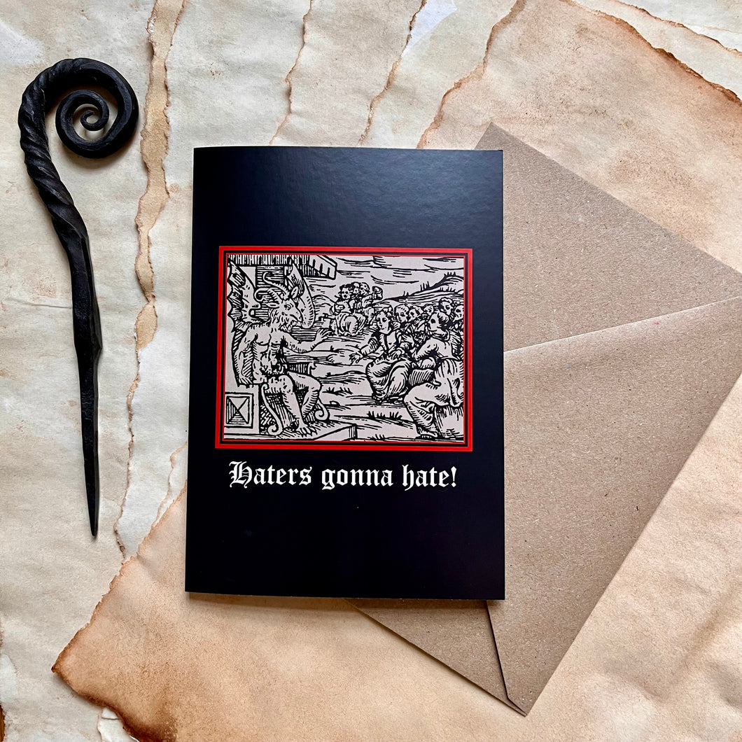 Photo of a black coloured card featuring a mediaeval woodcut artwork depicting witches approaching a demonic figure with a goat’s head and wings who sits on a throne and beckons to them. Beneath it the caption reads 'Haters gonna hate!'. Pictured with a brown envelope and black letter opener with a curved, spiral end.