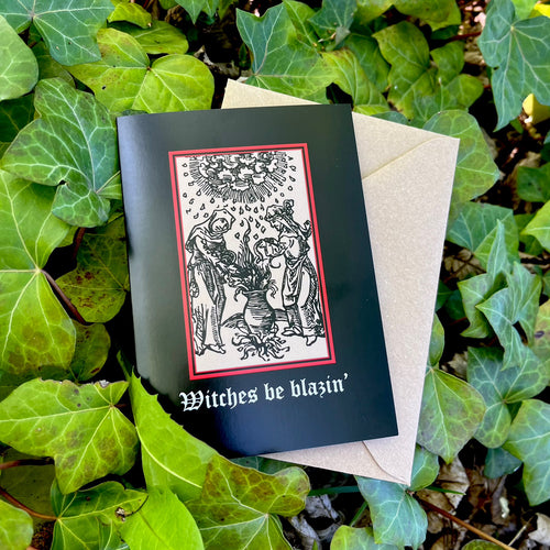 Photograph of a black card on a bed of ivy next to a brown envelope and leaves of multiple colours. The card features a mediaeval woodcut design of two women around a bubbling cauldron, as rain falls from a cloud overhead. Below in a black Gothic font are the words 
