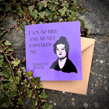 Load image into Gallery viewer, Photo of a purple greetings card on an ivy covered stone. The card depicts the quote &#39;I am no bird and no net ensnares me&#39; by Charlotte Bronte, with a portrait of her as a Goth.