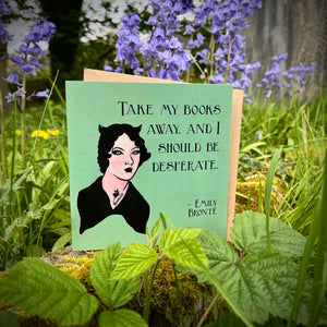 Photo of a green greetings card surrounded by nettles and bluebells. The card depicts the quote 'Take my books away, and I should be desperate' by Emily Bronte, with a portrait of her as a Goth.