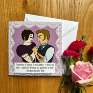 Greetings card with illustration of a couple in Victorian clothes in an embrace, with the man showing his country boy tattoos and the girl displaying her heavy metal tattoos. Card displayed beside flowers on a wooden surface.