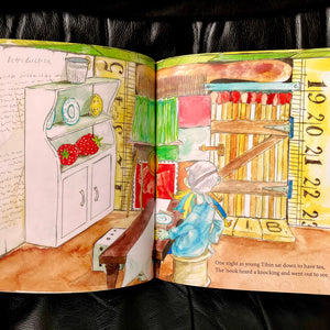 Photo of a double-page spread inside the book, showing a figure in a long blue top sitting at a wooden table. They are in a rustic kitchen with plates and gigantic strawberries on the shelf. They are looking towards the door. The caption reads “One night as young Tibin sat down to have tea, The ‘nook heard a knocking and went out to see.”