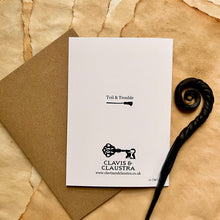 Load image into Gallery viewer, Photo of the back of the card, which is white and shows the Clavis &amp; Claustra logo along with the range name “Toil &amp; Trouble” and a broomstick. Pictured with a brown envelope and black letter opener with a curved, spiral end.