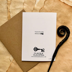 Photo of the back of the card, which is white and shows the Clavis & Claustra logo along with the range name “Toil & Trouble” and a broomstick. Pictured with a brown envelope and black letter opener with a curved, spiral end.